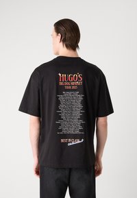 Černé bavlněné tričko s barevným grafickým potiskem na zadní straně, s textem "HUGO'S BIG DOG MINDSET TOUR 2025" a seznamem míst, lehce volný střih.