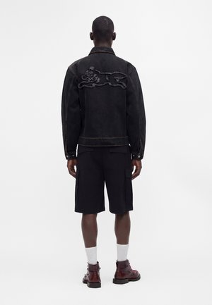 Mand stående med ryggen til, iført en sort denimjakke med broderet løbende hundemotiv på ryggen, sorte cargoshorts, hvide sokker og brune støvler.