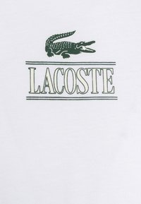 Balta medvilninė marškinėliai su žaliu krokodilo logotipu virš žodžio "LACOSTE" žaliais rašmenimis. Lygus paviršius, minimalus dizainas.