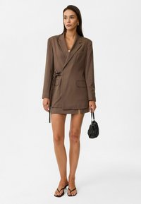 Bruine op maat gemaakte blazerjurk met een striktaille, voorzien van twee voorkeurszakken, een reverskraag en een gelaagde rok. Gestyled met zwarte accessoires.