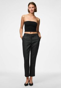 Črna brezramna majica s slim fit krojem; visoko pasne črne hlače z rahlim leskom; črne pointed balerinke; model pozira z rokami v žepih.