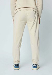 Pantalon de survêtement couleur crème avec une taille élastique, des poignets aux chevilles et une coupe ample, assorti à des baskets bleues et blanches.