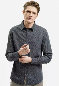 Mężczyzna poprawiający mankiet ciemnej koszuli z guzikami w mały biały wzór, stojący na tle jednolitego jasnego tła.