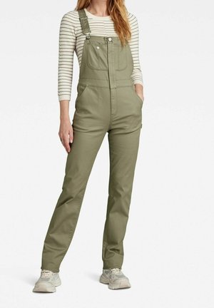 Kobieta w oliwkowo-zielonych ogrodniczkach z przednimi kieszeniami, w beżowo-czarną w paski bluzkę z długim rękawem i białych sneakersach, stojąca z rękami w kieszeniach.