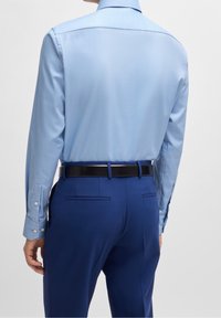 Jasnoniebieska koszula z długim rękawem o gładkiej fakturze, w połączeniu z ciemnoniebieskimi spodniami i czarnym paskiem. Widoczne guziki na mankietach.