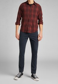 Camisa de cuadros roja y negra, combinada con jeans oscuros y zapatillas altas negras. La tela parece suave y tiene un ajuste casual.