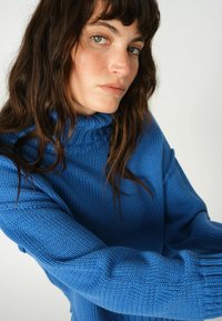 Plava pleta turtleneck pulover s rebrastom teksturom i dugim rukavima, opuštenog kroja i istaknutim detaljem ovratnika.