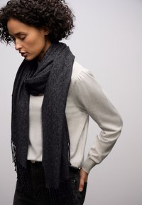 Écharpe noire à motif chevron avec un éclat métallique, bordée de franges, drapée autour d'un sweat-shirt gris clair.