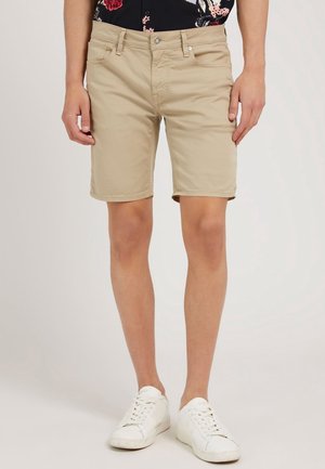 Beige Baumwollshorts mit lockerer Passform, Fünf-Taschen-Design und Reißverschluss, kombiniert mit weißen Sneakern.