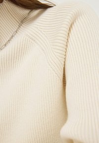 Cremfarbener gerippter Strickpullover mit hohem Kragen und sanft strukturierten Schultern. Verfügt über eine elegante silberne Kettenhalskette.