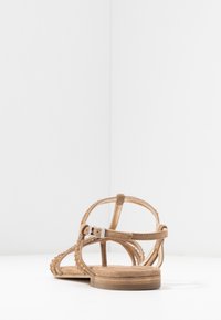 Sandale en suède beige avec des fines lanières, des clous ronds sur les côtés, une semelle plate et une fermeture à boucle réglable à la cheville.