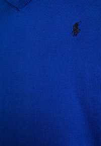 Blå stickad tröja med V-ringning och ribbat mönster. Har en liten svart logotyp på vänster bröst.