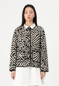 Quilted jakke med et dristig svart og kremfarget abstrakt mønster, knappelukking foran og to frontlommer. Hvit krage med avrundede kanter.