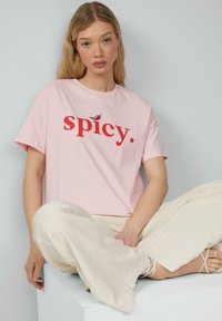 Rosa Baumwoll-T-Shirt mit dem Schriftzug "spicy." in fettem Rot, versehen mit einer kleinen Chili-Grafik, kombiniert mit cremefarbenen, weiten Hosen.