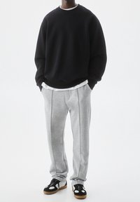 Man wearing black sweatshirt, white undershirt, gray sweatpants, and black-and-white sneakers standing with hands in pockets.