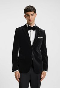 Zwart fluwelen tuxedojack met gepikte revers, single button sluiting en twee voorzakken. Witte zakdoek als accent. Glad texture.