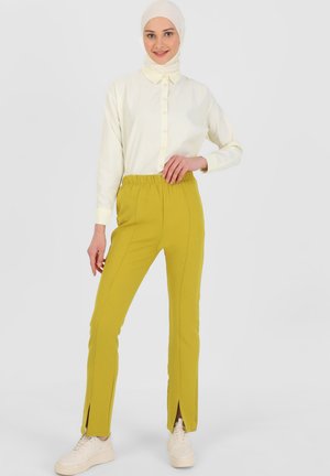 Camicia bianca con bottoni e maniche lunghe, abbinata a pantaloni gialli a gamba dritta con cintura elastica e spacchi laterali. Sneakers bianche.