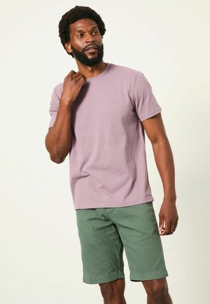 Bărbat cu barbă, purtând un tricou violet deschis și pantaloni scurți verzi, stând pe un fundal simplu și deschis, atingându-și gâtul.