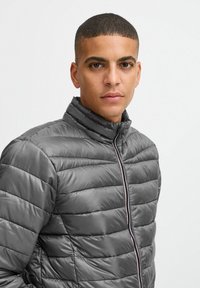 Grijze pufferjas met horizontaal quiltpatroon, hoge kraag en zwarte rits met een rode accent. Gladde, glanzende stoftextuur.
