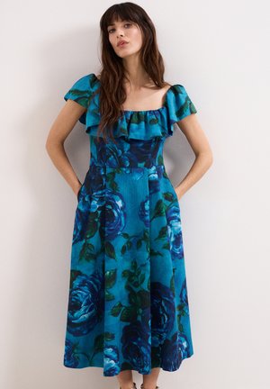 Femme en robe bleue fleurie à épaules dénudées avec volants, debout les mains dans les poches contre un mur clair uni.