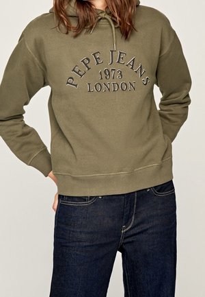 Osoba ubrana w oliwkowy kaptur Pepe Jeans London ze sznurkami i ciemnoniebieskie jeansy, stojąca z jedną ręką w kieszeni.