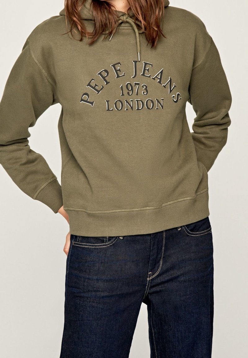 Personne portant un sweat à capuche Pepe Jeans London vert olive avec cordons et un jean bleu foncé, debout avec une main dans la poche.