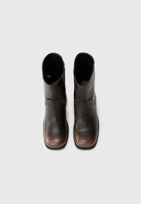 Par de botas de tornozelo de couro castanho escuro com bicos quadrados arredondados e saltos baixos, mostradas de cima sobre um fundo branco.