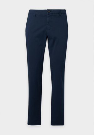 Homme portant un polo à manches longues gris clair, un pantalon slim bleu foncé, des chaussures noires, debout devant un fond clair uni.