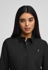 Camicia nera con bottoni e colletto strutturato, caratterizzata da un logo argento sul lato sinistro del petto e una texture di tessuto liscia e leggera.