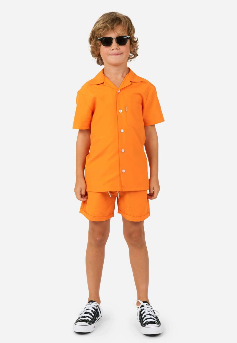 Camisa de manga corta de un brillante color naranja y pantalones cortos a juego, de material de algodón, con un bolsillo en el pecho. Gafas de sol negras y zapatillas de deporte en blanco y negro.