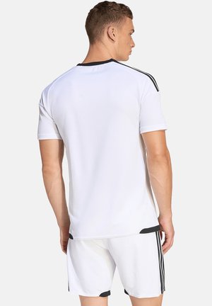 Mężczyzna ubrany w białe sportowe ubranie z czarnymi paskami na ramionach i bokach, stojący tyłem na jednolitym tle.