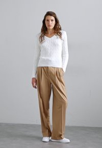 Pull en maille blanche à col en V associé à un pantalon beige décontracté. Le modèle porte des baskets blanches, sur un fond neutre.