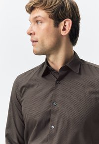 Brown patterned shirt with a button-down collar, featuring a textured fabric and blue buttons on the front, shown on a model's side profile.