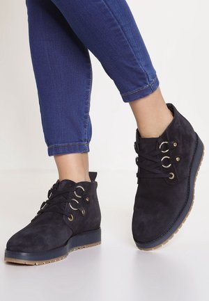 Botas de ante negras hasta el tobillo con puntera redonda, ojales dorados y cordones. Suela ligera marrón contrastante con agarre texturizado. Combinadas con unos vaqueros azules.