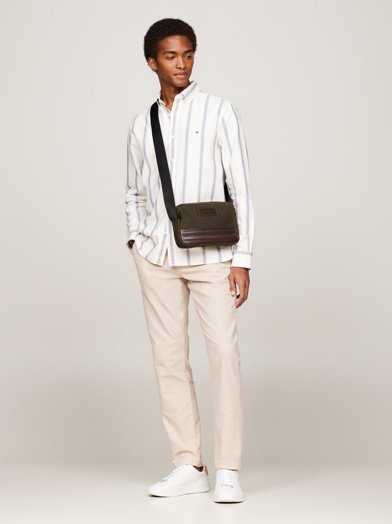 Mørk grønn budveske med en strukturert brun aksent, brukt over kroppen. Modell i hvitstripet skjorte og beige bukser, står i et nøytralt rom.