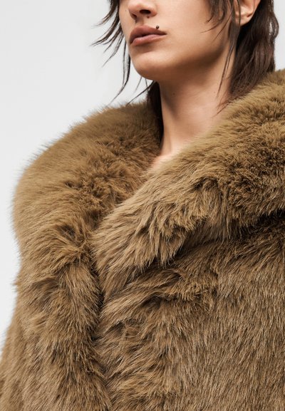 Άτομο που φορά ένα χοντρό, καφέ παλτό από συνθετική γούνα με ψηλό γιακά, εμφανίζοντας το κάτω μέρος του προσώπου και τον λαιμό, πάνω σε απλό φόντο.