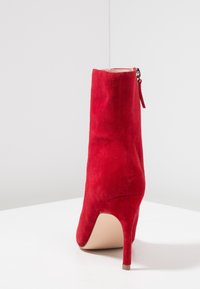 Botín tobillo de ante rojo con un alto tacón de aguja, detalle de cremallera lateral y textura suave; caña diseñada para un ajuste ceñido alrededor del tobillo.