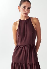 Nainen, jolla on selkähiukset, pukee ylleen hihattoman tumman luumunvärisen mekon, jossa on kerrostettu helma. Hän on asustautunut kultaisilla renkaankorviksilla ja seisoo valkoista taustaa vasten.