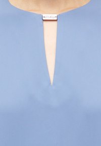 Gaiši zila blūze ar V veida kakla izgriezumu un sudraba emblemu detaļu pie apkakles. Izgatavota no gludas, vieglas auduma.