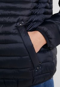 Veste rembourrée bleue marine avec des sections matelassées, une finition lisse, des poignets élastiques et une poche latérale à fermeture à bouton-pression.