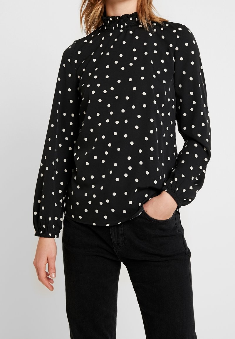 Blusa nera a maniche lunghe con pois bianchi, colletto alto con dettaglio arricciato e polsini elastici, realizzata in un tessuto leggero.