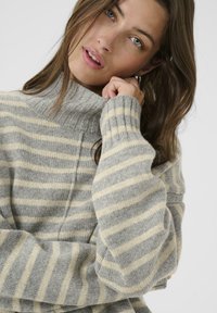 A young woman with long hair wears a striped sweater with high collar, playfully resting her chin on her hand, looking directly at the camera.