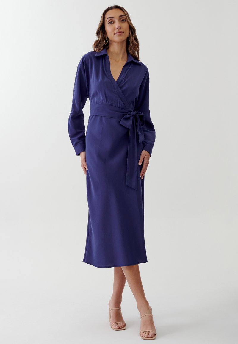 Abito avvolgente a maniche lunghe in un profondo blu navy, realizzato in tessuto morbido. Presenta una vita con cintura, scollatura a V e una lunghezza midi con una silhouette fluida.