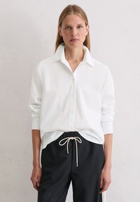 Biała koszula zapinana na guziki z kołnierzykiem, długimi rękawami i luźnym krojem, w połączeniu z czarnymi spodniami z elastycznym paskiem.