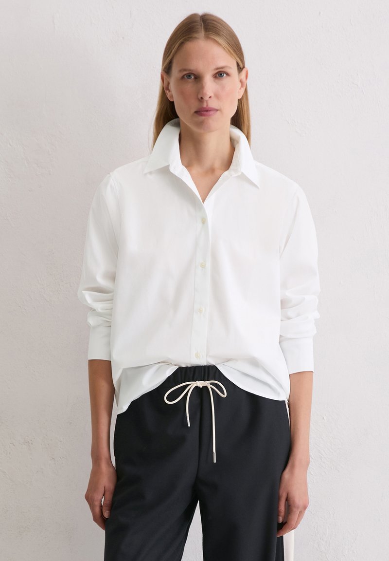 Biała koszula zapinana na guziki z kołnierzykiem, długimi rękawami i luźnym krojem, w połączeniu z czarnymi spodniami z elastycznym paskiem.