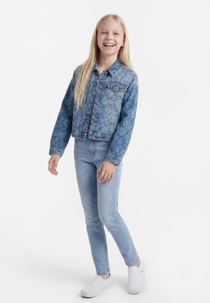 Lächendes Mädchen mit langen blonden Haaren, das eine Jeansjacke mit floralem Muster, hellblaue Jeans und weiße Slip-On-Schuhe trägt und vor weißem Hintergrund steht.
