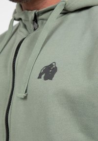 Groene rits-hoodie van zachte stof, met een zwart gorilla-logo op de borst en contrasterende zwarte ritsdetails.