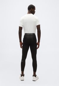 Άνδρας με άσπρο κοντομάνικο πουκάμισο, μαύρο κολάν Puma και άσπρα αθλητικά παπούτσια, στέκεται γυρισμένος με την πλάτη προς απλή ανοιχτόχρωμη επιφάνεια.