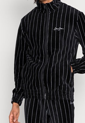 Veste zippée en velours noir avec des rayures verticales blanches. Dotée d'un col haut et de poignets élastiques, avec un petit logo brodé sur la poitrine.