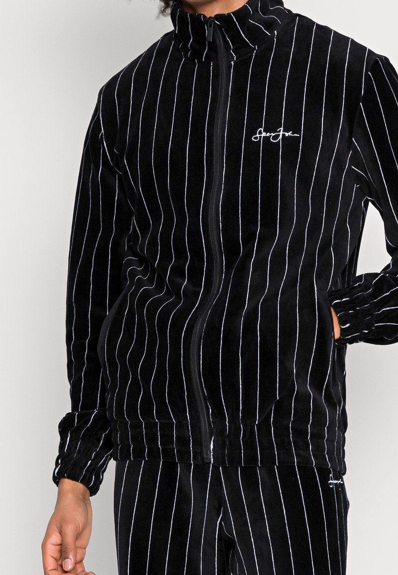 Veste zippée en velours noir avec des rayures verticales blanches. Dotée d'un col haut et de poignets élastiques, avec un petit logo brodé sur la poitrine.